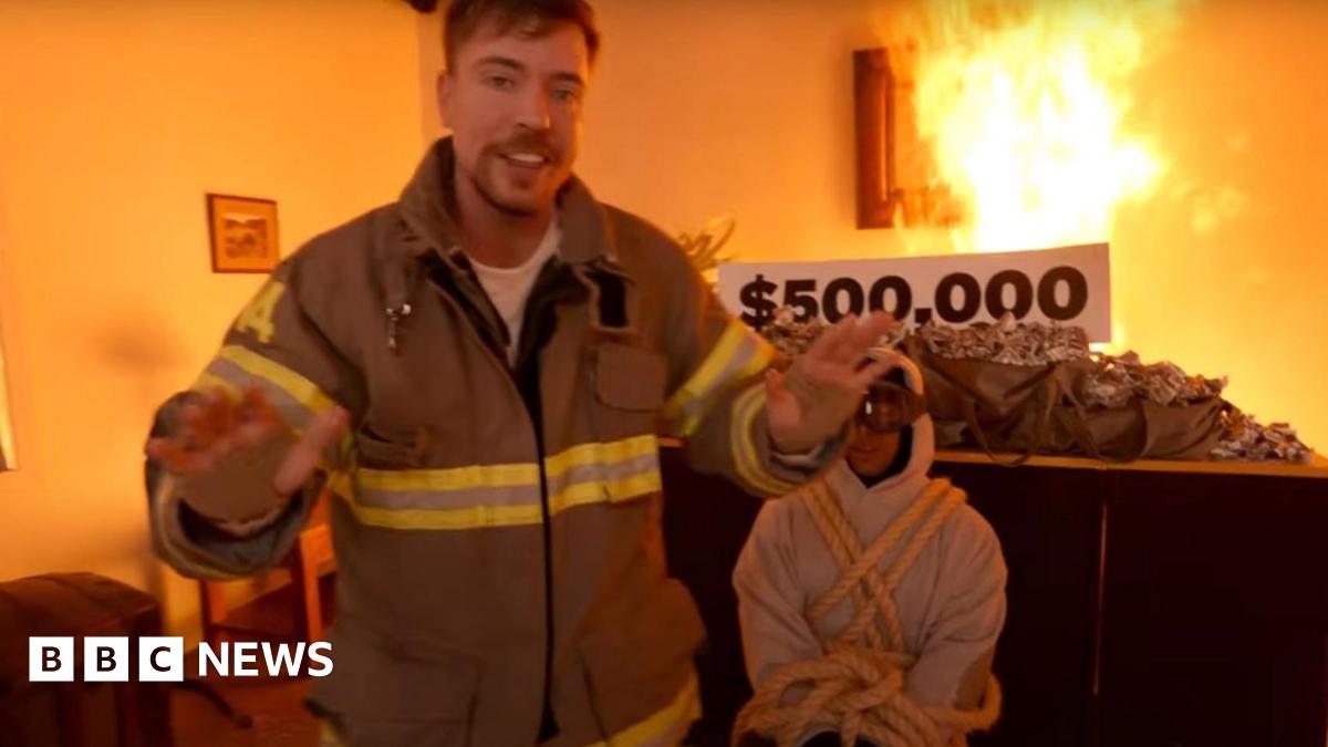 MrBeast, dressed in a firefighters jacket is standing in a burning room in front of a man in a white hoody, who is tied up. Behind both of them is a table piled high with sacks of dollar bills.