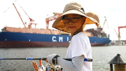 BBC A young man is smiling at the camera as he fishes on the dock. He is weating a sun hat, spectacles and a white t-shirt over a full-sleeved shirt. He has a watch on his left hand, wihich is holding the black fishing rod. 

Behind him is a long blue ship, with the letters C. M L on it in white. There are large cranes hovering over the vessel.     