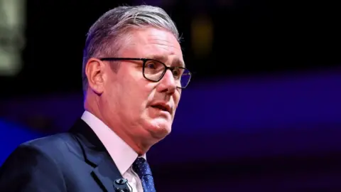 Reuters UK Prime Minister Keir Starmer sideways on in a navy blue jacket with a white shirt, navy tie with white polka dots and a small black mic on his lapel. He is also wearing black glasses.
