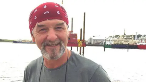 The Skipper magazine The late Kevin McCloskey pictured at a fishing harbour.  He is smiling at the camera.  He has a grey goatee beard and is wearing a red and white bandana and a grey t-shirt.  There is a body of water behind him and boats and buildings in the distance. 