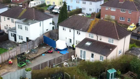 PA Media An aerial shot of a house on Blaise Walk in Sea Mills, Bristol. There is a white forensics tent in the back garden. The house is painted white with a brown roof. A police officer wearing a black uniform and a yellow high-vis jacket is standing in the garden. 