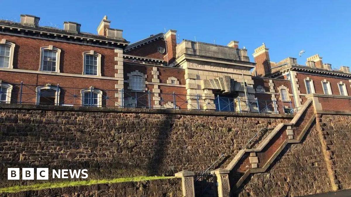 The exterior of HMP Exeter. It is a historic looking building of red brick with ornate granite block work and many chimneys. There is an inscription engraved in the stone above the entrance which reads: DEVON COUNTY PRISON. In the foreground is a stone wall with steps running through it.