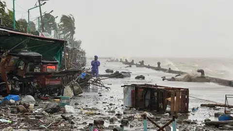 Getty Images A man in a blue raincoat stands amid rubble and debris on a sea shore that is being buffeted by wind, rain and waves. In the background are windswept palm trees.