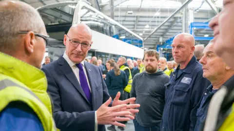 PA Media First Minister John Swinney, a man in his early 60s, surrounded by workers of a bus manufacturer