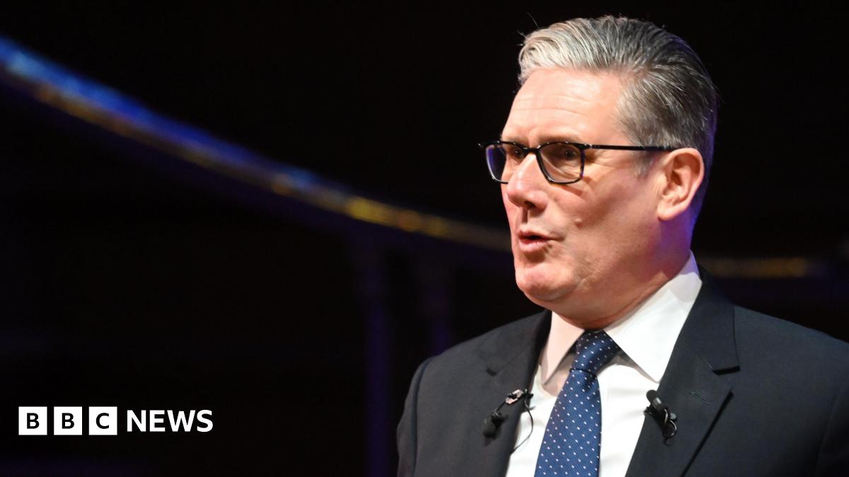 Sir Keir Starmer speaks while dressed in a dark suit, white shirt, and blue polka dot tie. He wears glasses and has short, neatly combed hair. Two microphones are clipped to his lapels. Behind him, a dark background features a softly lit curved structure.