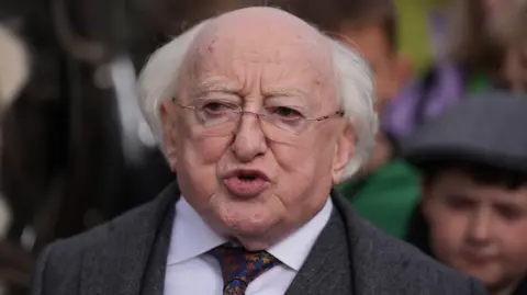 PA Media A close-up image of Michael D Higgins, who is attending the Ploughing Championships in County Offaly. President Higgins is an older man, who is balding with long white hair. He has frameless, rectangle glasses and is mid-speech. He is wearing a grey, woolen suit wth a white suit and blue and orange patterned tie.
