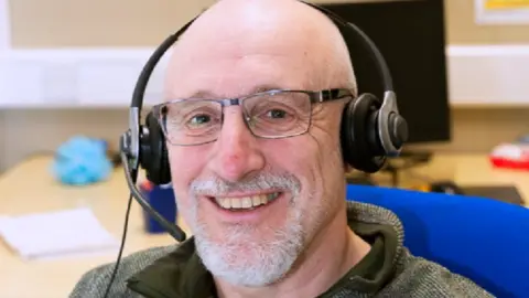 Samaritans James Watkins sits on a blue chair, wearing a phone headset, in front of an office desk. He is smiling, wearing a green top
