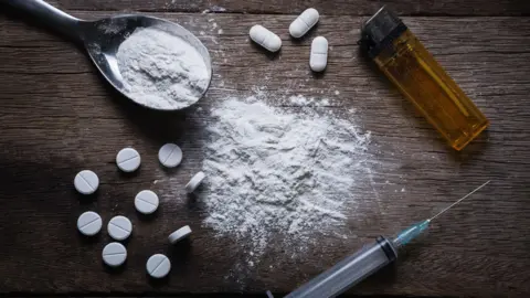 Getty Images A darkly-lit table with various drug-related paraphernalia. Several white pills are scattered alongside a hypodermic needle, a cigarette lighter and a spoon holding white power. There is more white powder on the table.