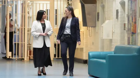 PA Media Shabana Mahmood visits HMP Bedford, alongside the prison's governor, Sarah Bott. The pair walk along a while corridor and in the background there is a prison officer holding open a white gate.