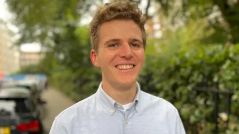 BBC/Fergus Walsh Jack May-Davis is smiling and is wearing a pale blue shirt. He is standing on a street with green hedges and a row of cars blurred in the background