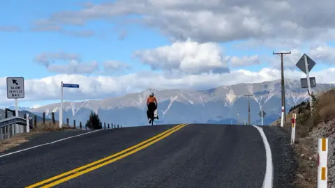 Duncan Covey A picture of a long road in New Zealand. There is a cyclist travelling on the left side of the road. There is a number of mountains and low clouds in the distance.