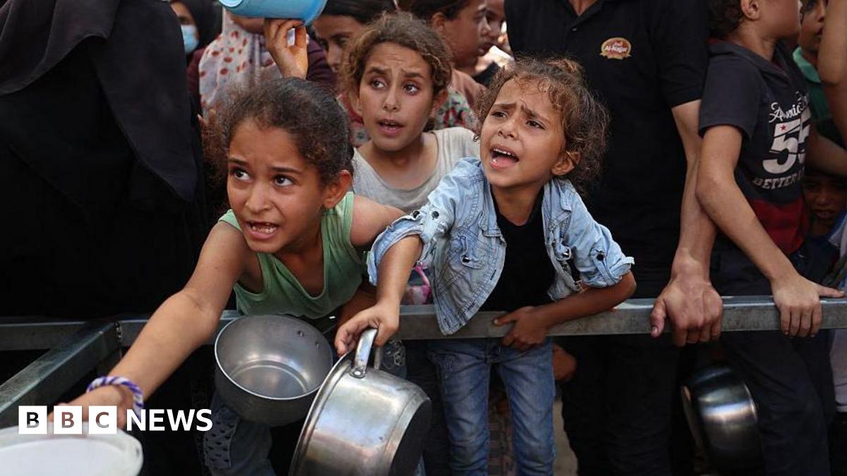 Palestinian children hold out pots at a charity kitchen in a refugee camp in the central Gaza Strip. They have pained looks on their faces and there is a crowd of people behind them