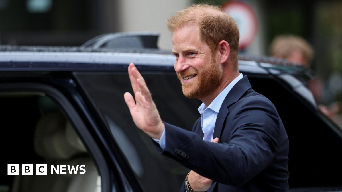 Prince Harry wearing a blue suit and blue shirt, waving as he steps into a black car.