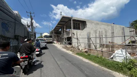 Ade Mardiyati A line of motorcycles waiting along a narrow road as a car is trying to squeeze past a large truck at a construction site.
