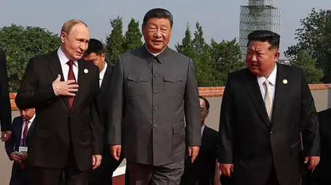 Getty Images Vladimir Putin, Xi Jinping and Kim Jong Un standing shoulder to shoulder, all of them in suits, with others in suits behind them. Putin is holding his right hand to his chest