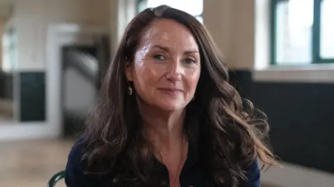A woman sitting in a bright room with wooden floors. She has long curly brown hair and is wearing a navy blue casual shirt. She smiles and looks at the camera.