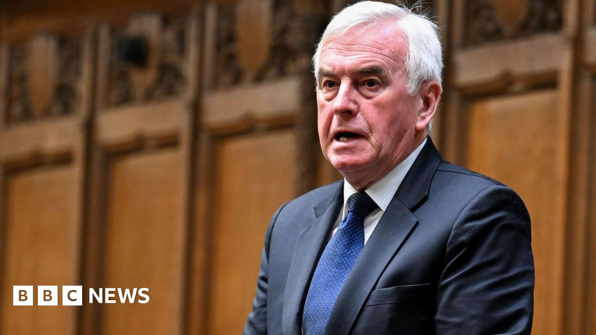 John McDonnell stands to deliver a speech in the House of Commons, dressed in a dark suit and blue tie. Behind him, the chamber’s traditional wooden panelling is visible.