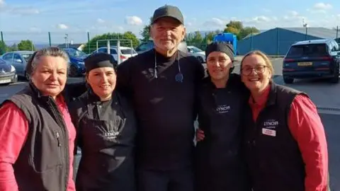 Lynch's Eurospar Greysteel Five people posing for a photograph outside a garage. 
One man standing in the middle of four women. All smiling at the camera. 
All of the women wear black work uniform. The man is pictured wearing black tracksuit bottoms a black jumper and a black hat. 
