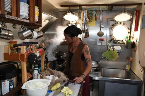 Matthias Berg/Klima Segler Giulia Baccosi cooking in a ship's kitchen. She has dark hair tied back and is wearing a brown waistcoat, read trousers and a black top. Her left arm has several tattoos and she is wearing a necklace. There is steam rising from a large pan which she is holding a spoon above. In front of her is a chopping board and a large knife with a potato-like vegetable which has been chopped and another whole one. There is a basket of large white eggs, plates, jugs and other equipment hanging up to her side and behind her. 