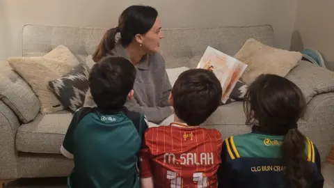 Karen Cullen Karen Cullen is holding a book and reading to her children while sat on a sofa.
Her three children (L-R) Ben, Brandon and Emily. They are wearing GAA and football jerseys and listening intently to their mum. 