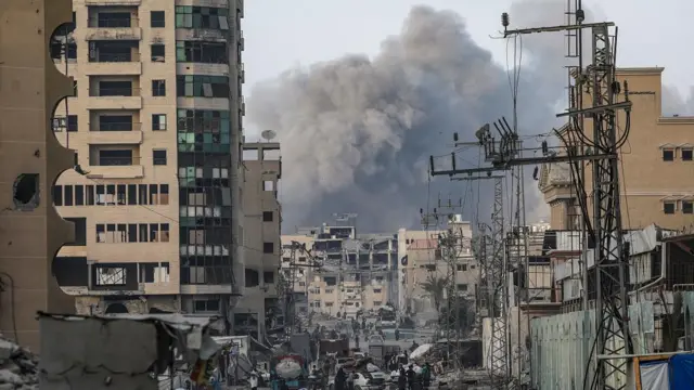 A large plume of smoke rises over Gaza city, where people walk below damaged buildings