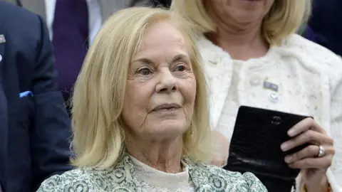 Rebecca Naden/PA A woman with blonde hair and a green floral jacket is standing with a crowd of people behind her. She is looking into the distance.