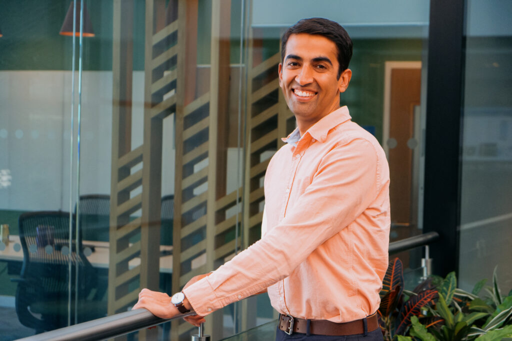 A portrait of a black-haired man in a peach-colored oxford shirt. He's holding the handrail of a walkway and looking over his left shoulder at the camera.