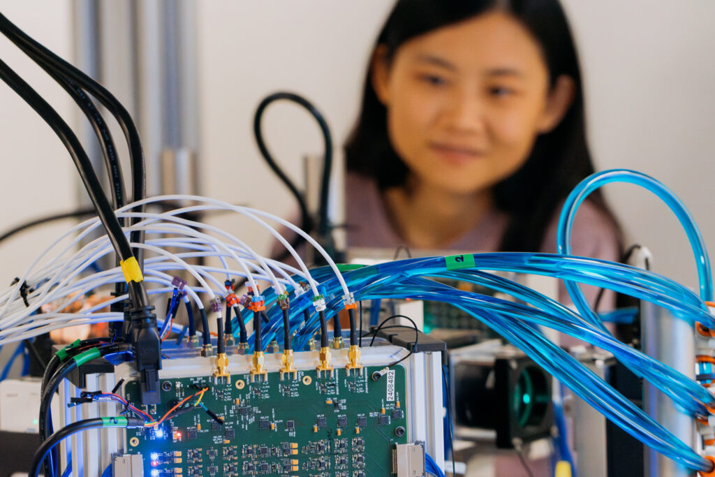 A woman with long black hair is out of focus behind a device made of many wires and glowing with lights.