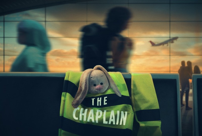 A stuffed bunny resting on a vibrant green vest labelled 'THE CHAPLAIN' in an airport setting, with blurred passengers and an airplane taking off in the background during sunset.