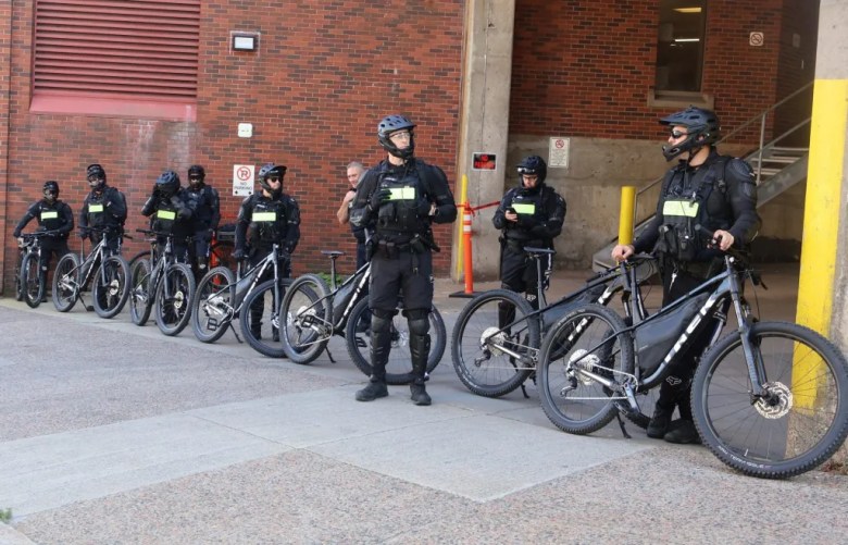 Security personnel dressed in black and wearing helmets are blocking the entrance to a stadium, each holding a bike.