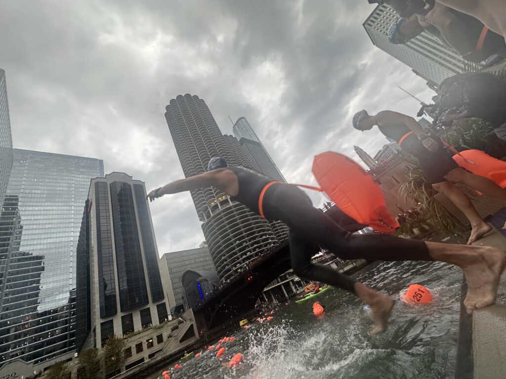 People are wearing blue swim caps and neon orange buoys to be more visible. The image shows a person mid-jump as another splashes in, skyscrapers looming over them