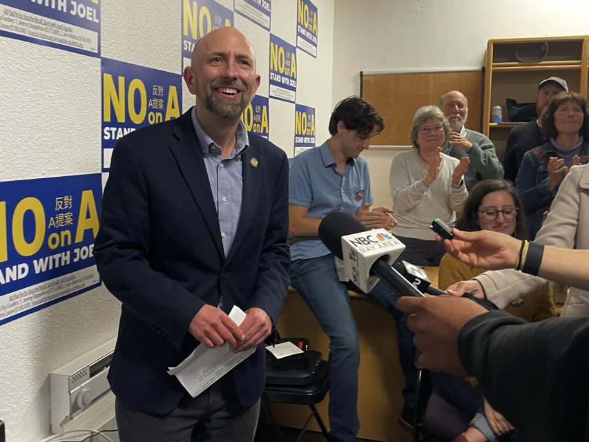 A man in a suit smiles while speaking to reporters at an indoor event; "NO on A" signs cover the wall, and people sit and stand behind him, some clapping.