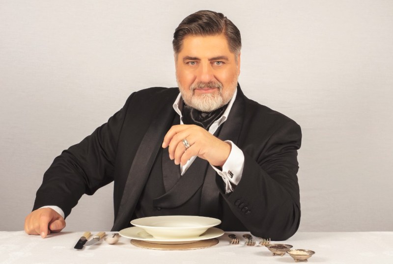 Man in formal attire seated at a dining table, holding a wine glass, with an elegant white plate and cutlery set in front of him, exuding a sophisticated dining atmosphere.