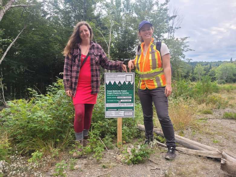 A woman with long brown hair, wearing a checkered red and black flannel jacket over a red dress, with red leggings and thick grey socks and hiking boots, stands to the left of a green and white sign for a "Forest Herbicide Project" that is on a post stuck in the ground. She points to an empty white square in the upper right hand corner of the sign. A woman on the right side of the sign, wearing a blue baseball cap, and orange and yellow vest over a beige t-shirt, and black pants and running shoes, points to the same empty white box on the sign. The backdrop is a previous clearcut area on the right, and some taller intact woodlands to the left behind them.