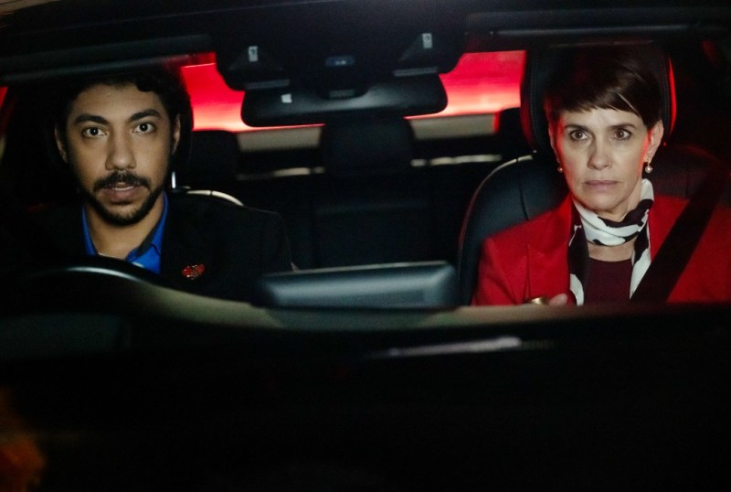 A man and a woman sitting in a car at night, both looking serious. The man has curly hair and is wearing a black blazer with a blue shirt, while the woman is dressed in a red jacket and a striped scarf. The car's interior is dimly lit, with a red glow visible outside.