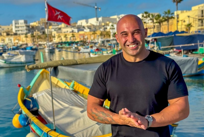 Smiling man in a black t-shirt standing by a picturesque harbour with traditional fishing boats in Malta, showcasing vibrant waterfront buildings and a clear blue sky.