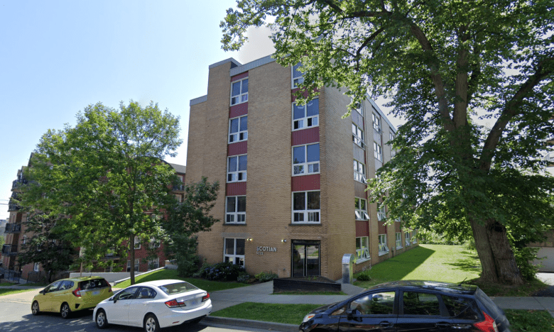 A yellowish five-storey apartment building is flanked by two large trees.