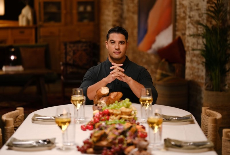 A man seated at a beautifully set dining table featuring a variety of gourmet food, including fruits, bread, and meats, with glasses of white wine in the background. The setting is warmly lit, creating an inviting atmosphere for a meal.
