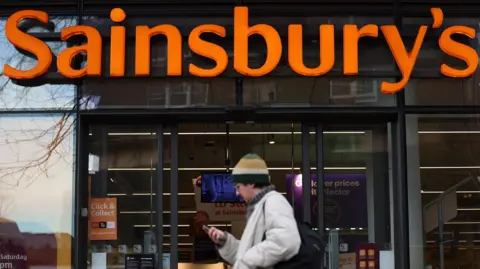 EPA The entrance of a Sainsbury's supermarket with a large orange "Sainsbury's" sign above glass doors. A person wearing a beige coat, green and beige beanie, and carrying a black backpack walks past while holding a phone. Inside the store, shelves and signage are visible, including a "Click & Collect" sign on the left.