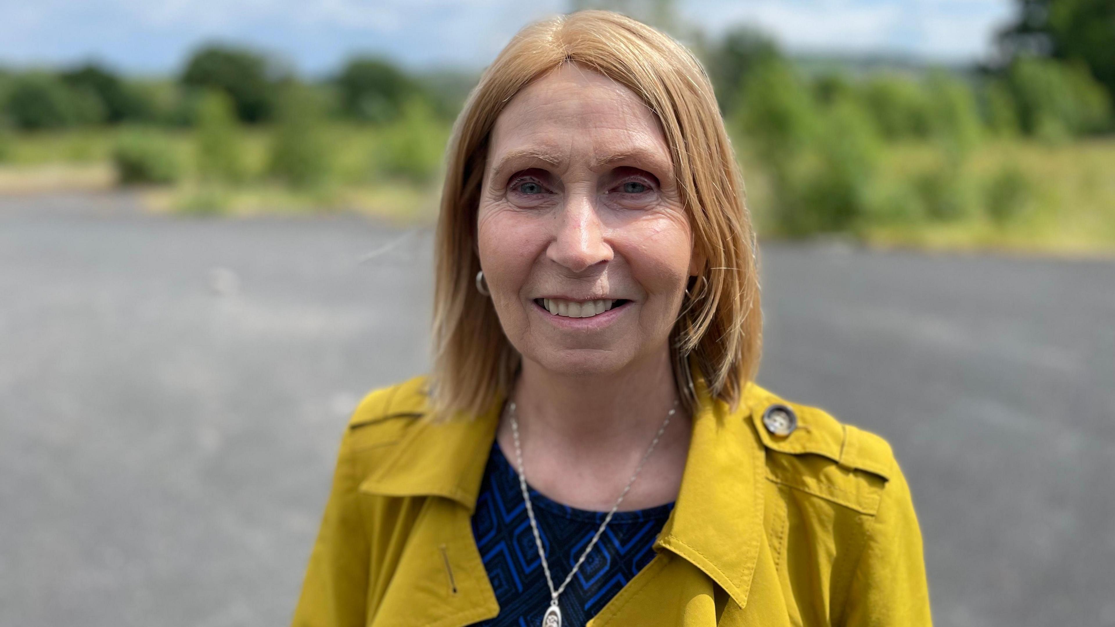 Linda Ervine smiling at the camera. She has light hair and is wearing a yellow jacket.
