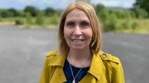 Linda Ervine smiling at the camera. She has light hair and is wearing a yellow jacket.