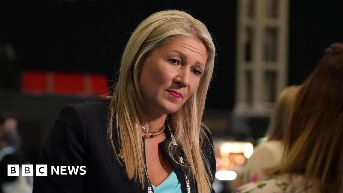 Laura Anne Jones speaking to an individual off camera, wearing a black jacket and blue top.