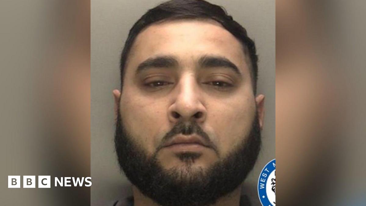 A police mugshot of a man with short black hair and a large black beard.