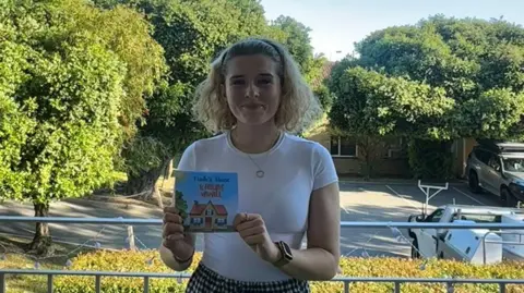 Síofra Harkin Síofra Harkin is holding a copy of her book on a balcony.
She has long blonde hair tied back with a black headband.
She is wearing a white top and a checked skirt.
