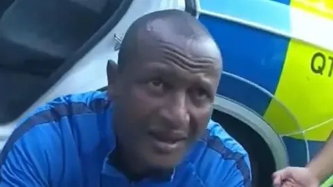 Essex Police Hadush Kebatu has short black hair and is wearing a blue sports top. He is crouched in front of a police car and has his eyebrows raised.