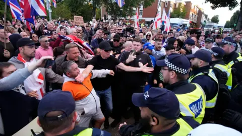 PA Media A large crowd of demonstrators, some of whom are waving flags and signs, get close to a line of police officers during a protest march in Epping.