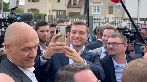 Jordan Bardella takes a selfie in the middle of a crowd of journalists and supporters at a rally