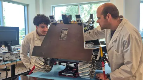 Engineers Misha Hartmann and Adam Abdulahad work on a prototype of the rover at the Canadensys headquarters. Both wear white lab coats and are working on the vehicle, which is siting on a table, and has metal wheels and visible wires.