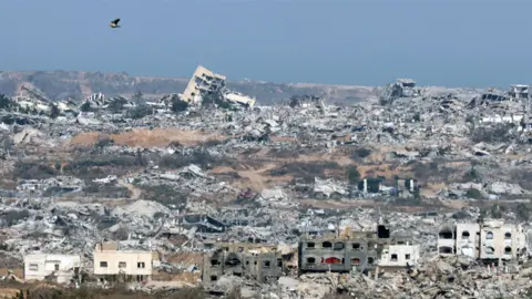 AFP via Getty Images The image shows a vast area of destruction with numerous collapsed and damaged buildings. Rubble and debris are scattered across the landscape, indicating extensive devastation. In the foreground, there are partially standing structures with visible damage. A bird is flying in the sky above the ruins. The background features more destroyed buildings extending to the horizon under a clear blue sky.