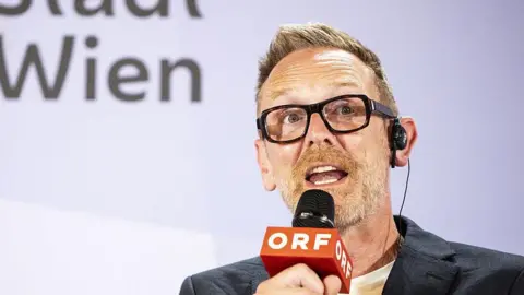 Getty Images Director of The Eurovision Song Contest (ESC) Martin Green speaks during a press conference for the announcement of the host city of the upcoming Eurovision Song Contest on August 20, 2025 in Vienna. He has short light brown hair and beard, wearing glasses and an ear piece, holding a microphone with 'ORF' on it. He is wearing a black blazer and white t-shirt.
